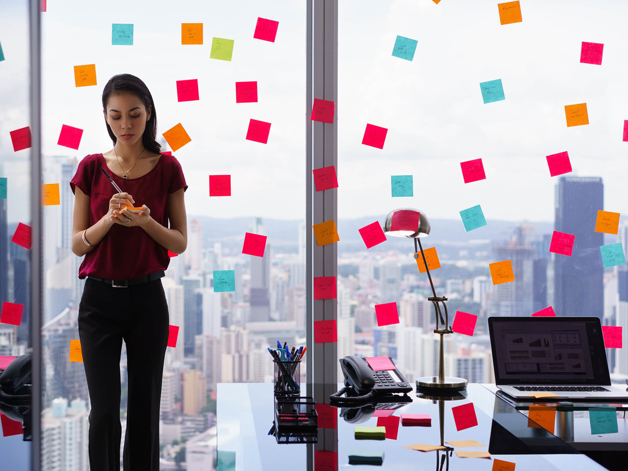 Mixed race secretary working in modern office in skyscraper  writing and sticking adhesive notes with tasks on window  
