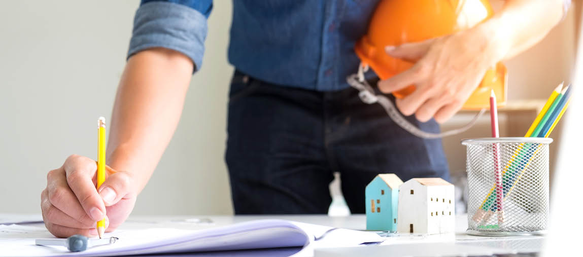 Architects are using drawing pencils,On the other hand holds a safety hat,On the writing desk there are model houses, laptop and house plans.