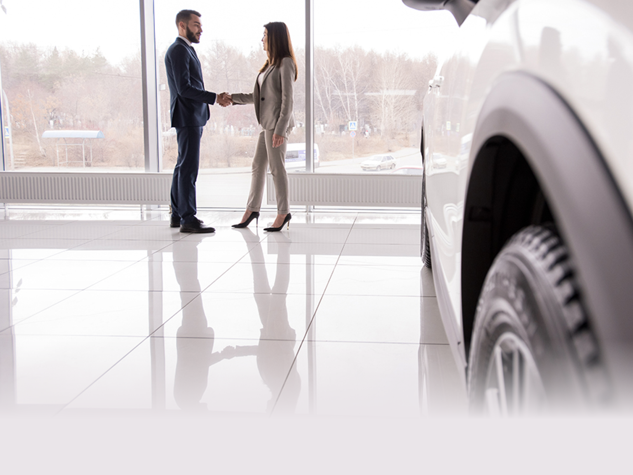 Wide angle portrait of car salesman shaking hands with woman buying new car in dealership showroom, car wheel in foreground, copy space
